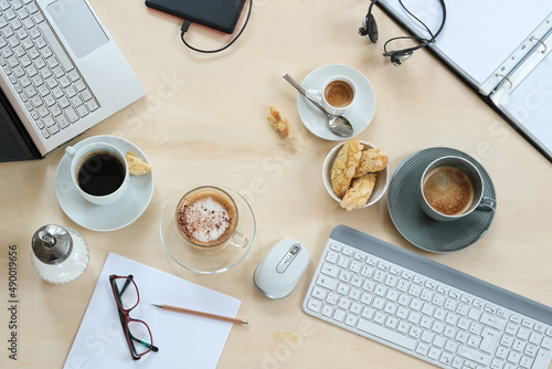 Meeting table seen from above with coffee cups and cookies, laptop, keyboard, folder and papers on a light wooden background, nobody