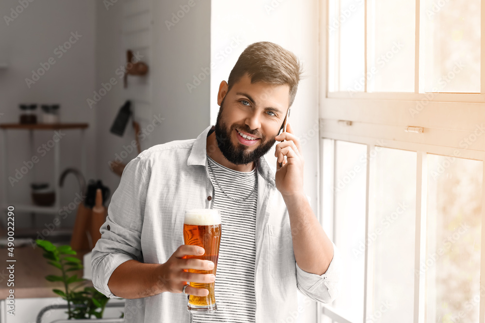 Handsome bearded man with glass of beer talking by mobile phone in kitchen Handsome bearded man with glass of beer talking by mobile phone in kitchen