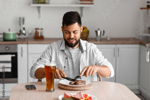 Handsome bearded man eating sausages at table in kitchen