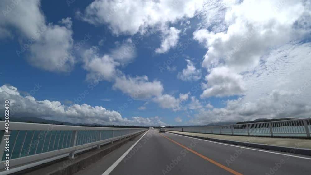 OKINAWA, JAPAN - AUG 2021 : Driving around Kouri Island bridge in sunny summer day. Wide camera, point of view (POV), seaside road driving. Holiday, vacation and resort concept. OKINAWA, JAPAN - AUG 2021 : Driving around Kouri Island bridge in sunny summer day. Wide camera, point of view (POV), seaside road driving. Holiday, vacation and resort concept.