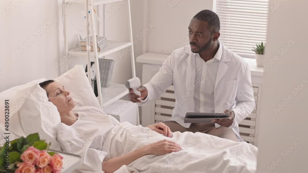 Medium slowmo of African American doctor using contactless infrared thermometer and tablet pc measuring temperature of female patient sleeping in hospital bed recovering from therapy or surgery Medium slowmo of African American doctor using contactless infrared thermometer and tablet pc measuring temperature of female patient sleeping in hospital bed recovering from therapy or surgery