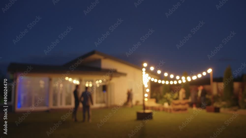 Blurred image of People stand in groups and talk in the courtyard near the cottage where the party with colorful lightmusic takes place Blurred image of People stand in groups and talk in the courtyard near the cottage where the party with colorful lightmusic takes place