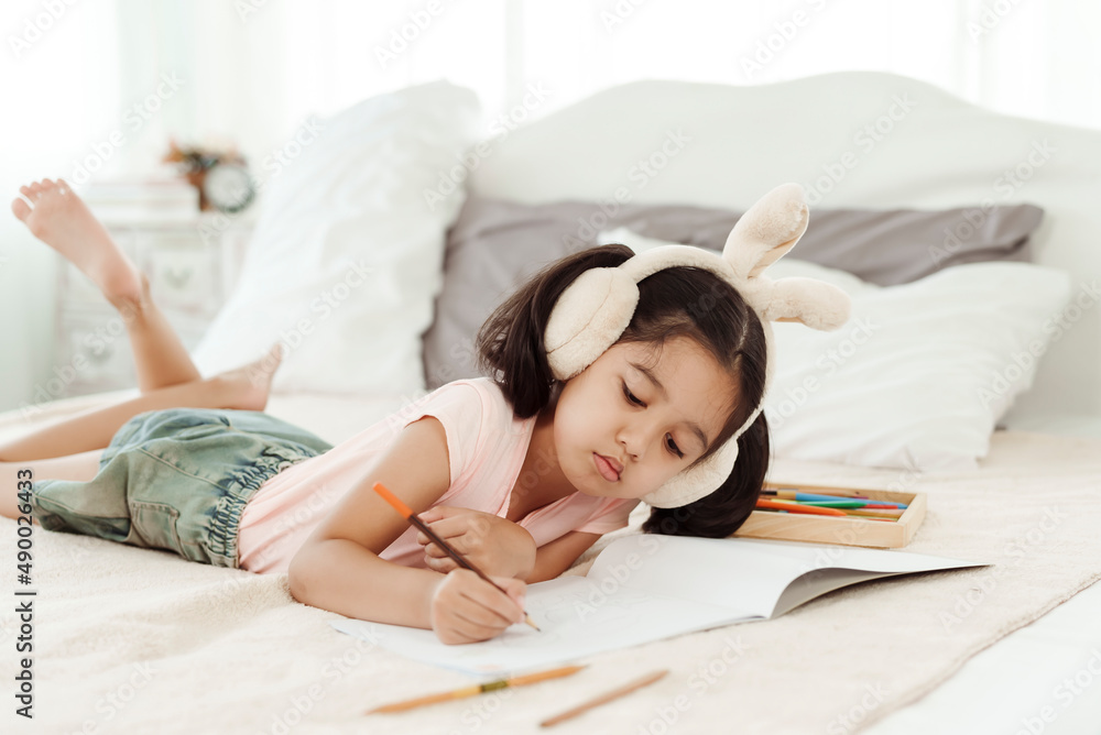 Cute little girl having fun in cute kindergarten age. Use colored pencils to draw a picture on the paper. Happily put the rabbit headphones on the bed. Concept family and happy learning Cute little girl having fun in cute kindergarten age. Use colored pencils to draw a picture on the paper. Happily put the rabbit headphones on the bed. Concept family and happy learning
