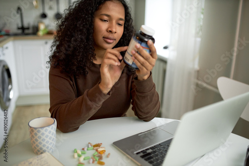 Charming black ethnicity female of 30s sitting at kitchen table in front of opened laptop, cup and vitamin pills, holding bottle of food supplements, reading suggested usage and warnings photo