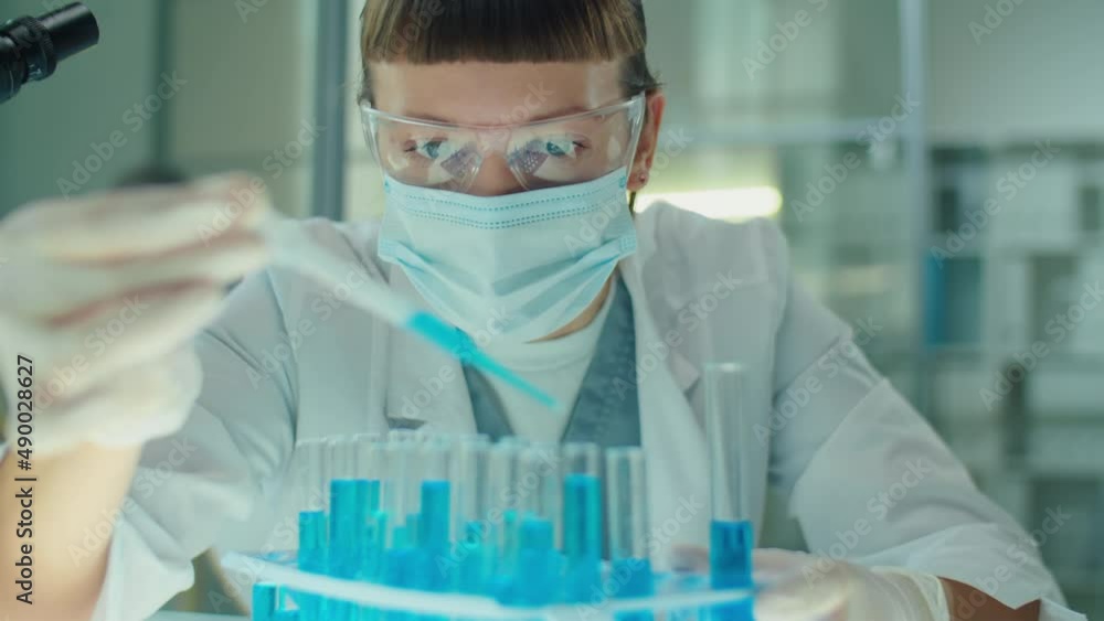 Female microbiology scientist in protective face mask, glasses and gloves pouring blue chemical into test tubes with pipette in laboratory while working on vaccine against covid Female microbiology scientist in protective face mask, glasses and gloves pouring blue chemical into test tubes with pipette in laboratory while working on vaccine against covid