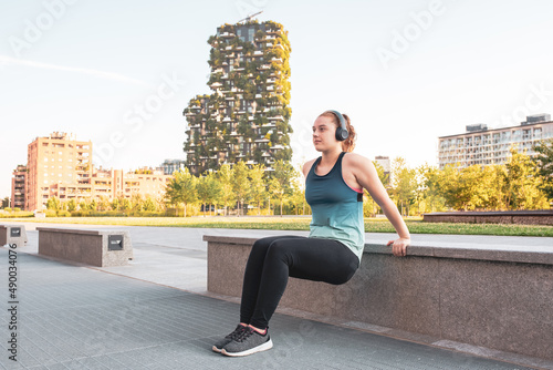 a young woman doing exercise outdoors in a public park, girl of the generation z sports prepares for the summer costume test, copy space, urban skyscrapers background, blu and black sportwear