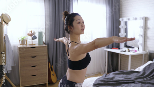 waist up slender girl wearing sportswear bra practicing warrior II pose or virabhadra in her bedchamber. slowly stretching arms parallel to the floor and gazing into distance with concentration. photo