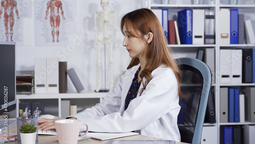 portrait asian female doctor sitting in the office is looking up information on her computer and transcribing on notebook with pen.