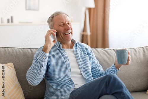 Cheerful mature man talking on cellphone sitting on couch