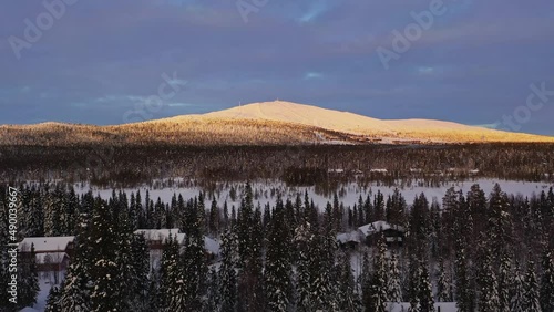 Aerial view overlooking cabins in middle of snowy trees, Yllastunturi fell, sunrise background, in Yllas, Lapland - tracking, drone shot photo