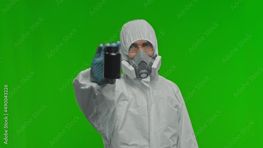 Scientist points with his index finger to a bottle of pills. He is wearing a respirator, glasses and a protective suit. Scientist points with his index finger to a bottle of pills. He is wearing a respirator, glasses and a protective suit.