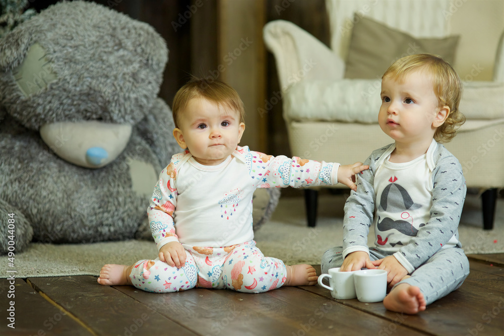 Two small children aged one year in pajamas are playing with cups and a kettle. Home environment. Two small children aged one year in pajamas are playing with cups and a kettle. Home environment.