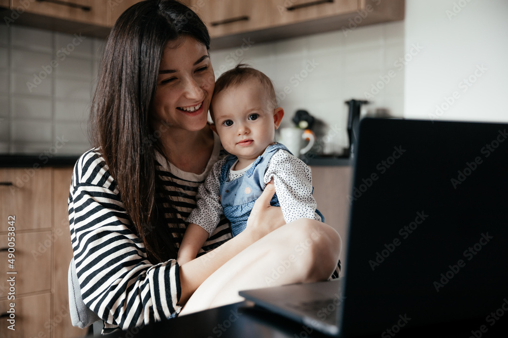 a young mother with many children working on a laptop sitting in the kitchen with a baby in her arms while the eldest daughter helps with cooking and cleaning in the kitchen a young mother with many children working on a laptop sitting in the kitchen with a baby in her arms while the eldest daughter helps with cooking and cleaning in the kitchen