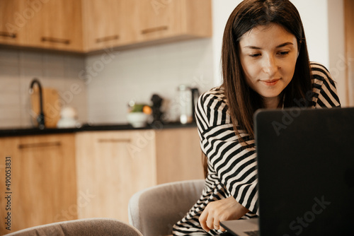 Young female freelancer dressed in home clothes working on laptop sitting at the table in the kitchen. Stylish girl on workplace at the desk. Distance learning online education