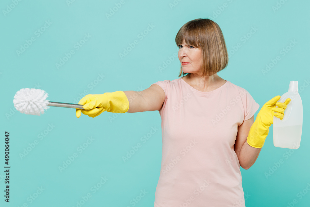 Elderly strict housewife woman 50s in t-shirt gloves hold toilet brush sponge detergent chemical bottle clenser isolated on plain pastel light blue background Housekeeping cleaning tidying up concept Elderly strict housewife woman 50s in t-shirt gloves hold toilet brush sponge detergent chemical bottle clenser isolated on plain pastel light blue background Housekeeping cleaning tidying up concept