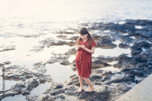 Cute long-haired European kid girl in a dress on the sea coast. A teenager child walks along a deserted embankment, summer vacation and children's emotions