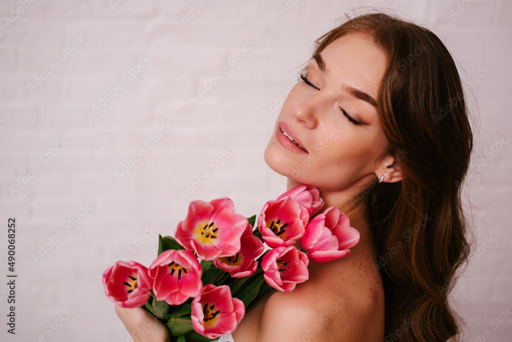 Gentle portrait of a beautiful woman with red long hair, closed eyes, bare shoulders on a white background. Girl with a pink bouquet of tulips.
