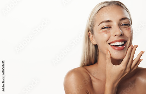 Body and skin care. Happy blond woman with white smile, perfect healthy teeth, touching clean, glowing facial skin, using under eye patches, scrub, standing over white background