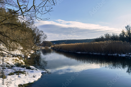 Evening on a forest river, a mirror-like surface of water and a clear sky, grass and trees on the banks, snow in some places, a beautiful winter landscape