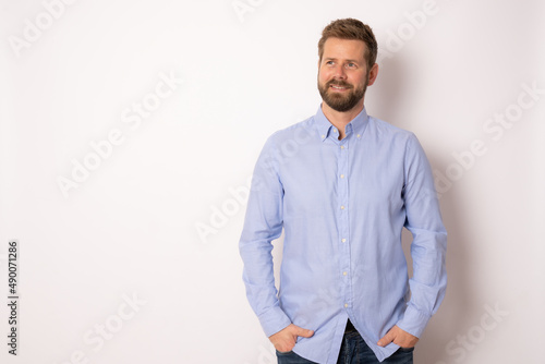 Portrait of charming successful young man in blue shirt, smiling broadly with self-assured expression while holding hands in pockets over white background. Business concept.