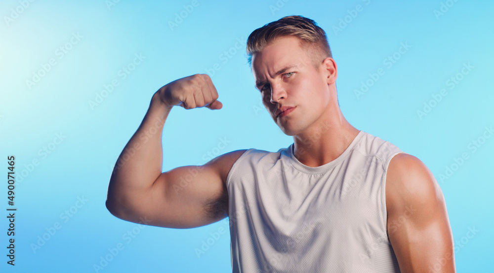 Welcome to the gun show. Studio portrait of a handsome young man flexing his bicep against a blue background. Welcome to the gun show. Studio portrait of a handsome young man flexing his bicep against a blue background.