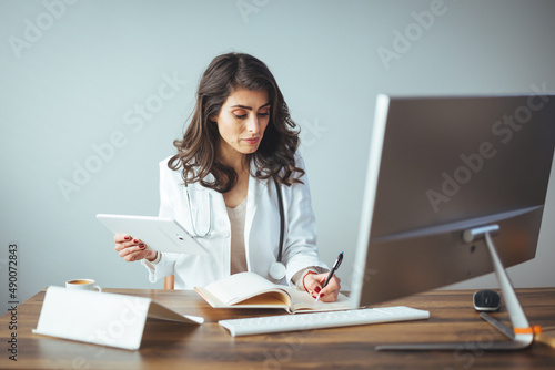 Female doctor using a digital tablet at her desk in the clinic. Female doctor working on her tablet pc in her office. Mid adult female doctor reviews patient records on desktop PC photo