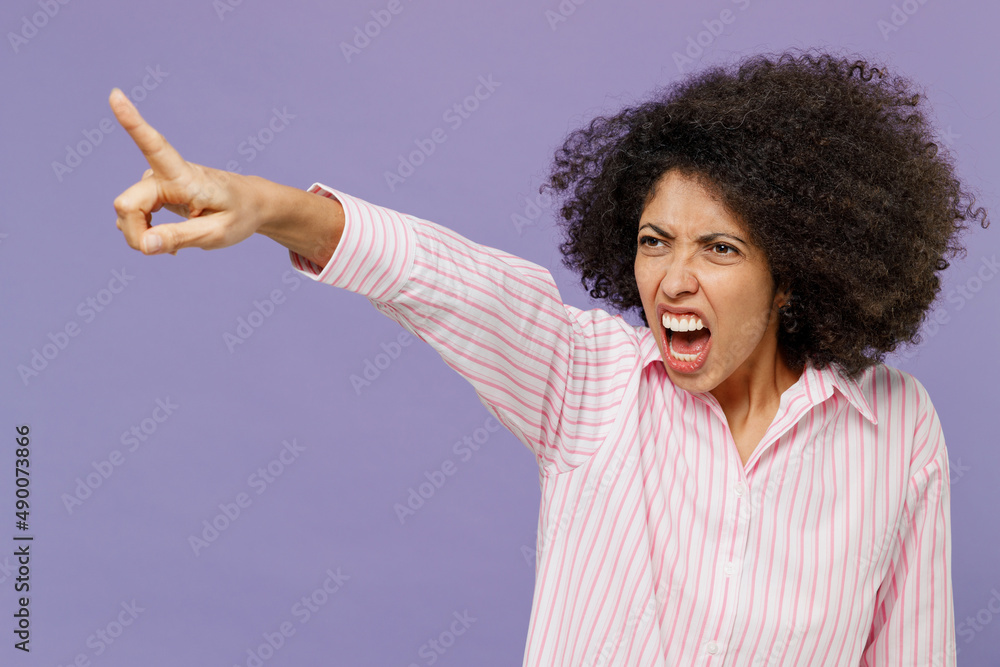 Young furious angry sad indignant woman of African American ethnicity 20s wearing pink striped shirt point index finger aside scream isolated on plain pastel light purple background studio portrait. Young furious angry sad indignant woman of African American ethnicity 20s wearing pink striped shirt point index finger aside scream isolated on plain pastel light purple background studio portrait.