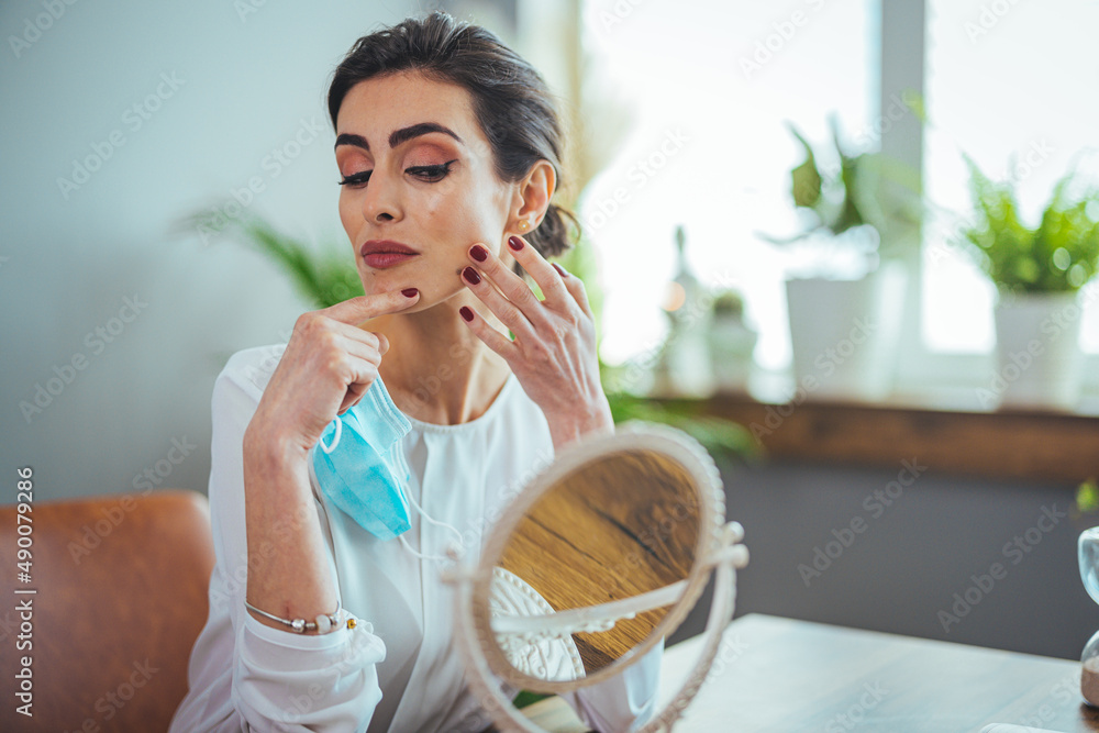 Young beautiful woman looking herself in the mirror at home. Worried about acne caused by wearing a mask. Maskne. Problems with acne. Young beautiful woman looking herself in the mirror at home. Worried about acne caused by wearing a mask. Maskne. Problems with acne.