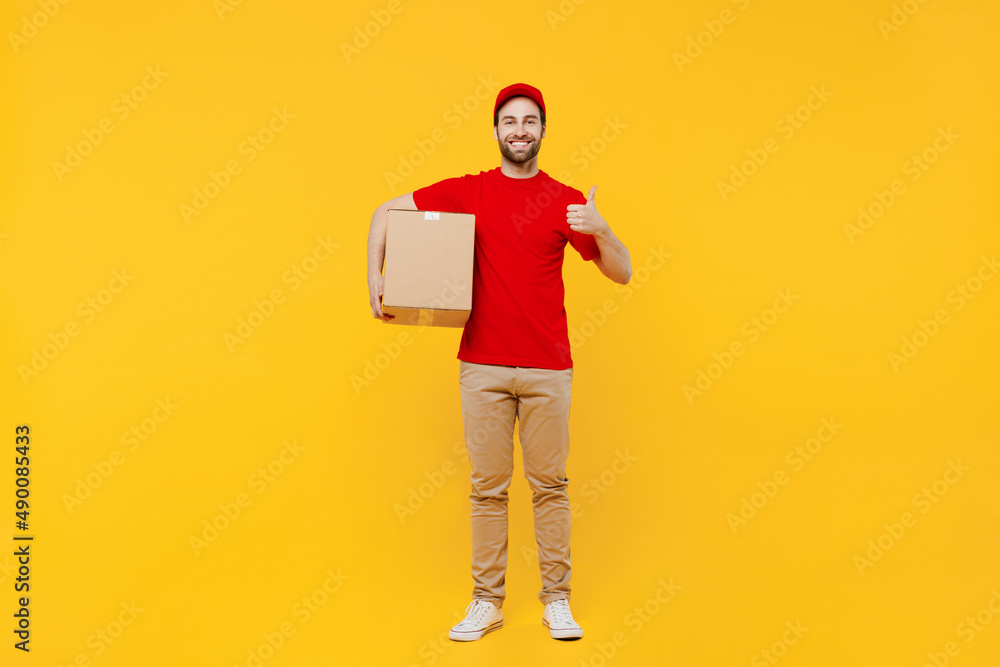 Full body smiling fun happy delivery guy employee man in red cap T-shirt uniform workwear work as dealer courier hold cardboard box show thumb up isolated on plain yellow background studio portrait Full body smiling fun happy delivery guy employee man in red cap T-shirt uniform workwear work as dealer courier hold cardboard box show thumb up isolated on plain yellow background studio portrait