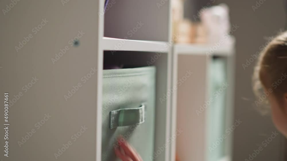 Pretty blonde little girl looks inside large fabric containers on wooden rack in light children room at home close side view slow motion Pretty blonde little girl looks inside large fabric containers on wooden rack in light children room at home close side view slow motion