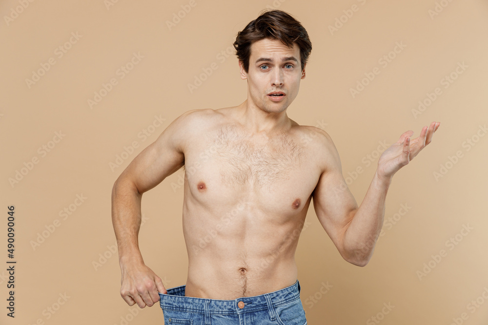 Attractive young man 20s with naked torso showing loose pants spread hand isolated on pastel pastel beige color background Body care healthcare fitness sport bodybuilding diet loosing weight concept.