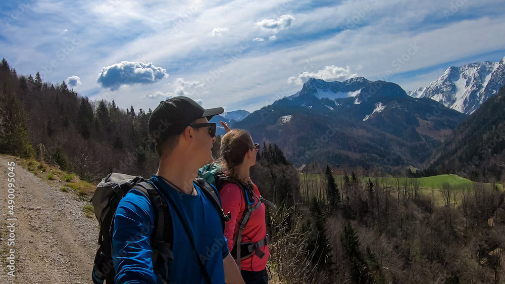 Couple with backpack and scenic view of snow capped mountain peaks of Karawanks near Sinacher Gupf in Carinthia, Austria. Mount Hochstuhl (Stol) visible through forest in spring. Rosental sunny day