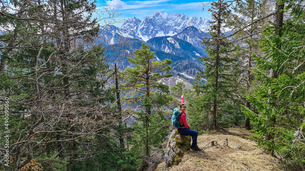 Obraz premium Woman with backpack sitting on a rock with scenic view of snow capped mountain peaks of Karawanks near Sinacher Gupf in Carinthia, Austria. Mount Wertatscha is visible through dense forest in spring
