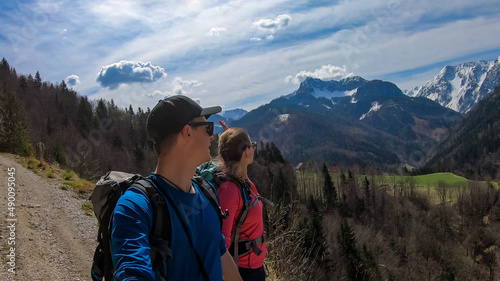 Couple with backpack and scenic view of snow capped mountain peaks of Karawanks near Sinacher Gupf in Carinthia, Austria. Mount Hochstuhl (Stol) visible through forest in spring. Rosental sunny day