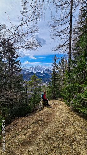 Woman with backpack sitting on a rock with scenic view of snow capped mountain peaks of Karawanks near Sinacher Gupf in Carinthia, Austria. Mount Wertatscha is visible through dense forest in spring