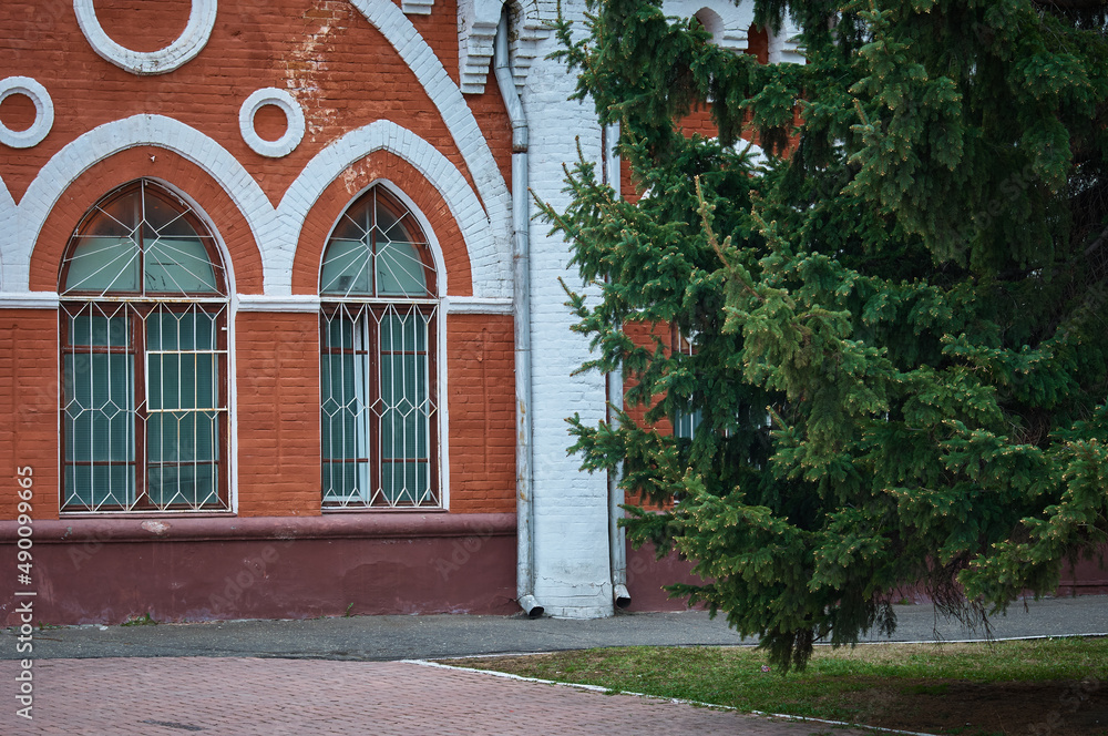 The facade of a historical building made of red brick in the pseudo-Gothic style of architecture. Fluffy branches of an evergreen spruce. Former malls. 