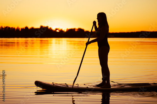 Silhouette of middle aged Caucasian woman standing on sup board with oar in hands on lake in evening with magnificent sunset in background. Active lifestyle.