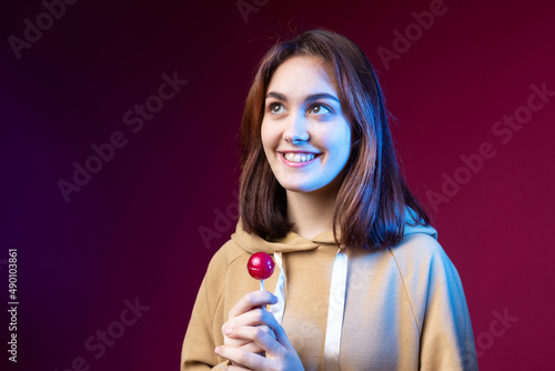 Portrait of a woman in a hoodie  girls licking a red round lollipop with beautiful makeup on a purple trendy background in the studio.