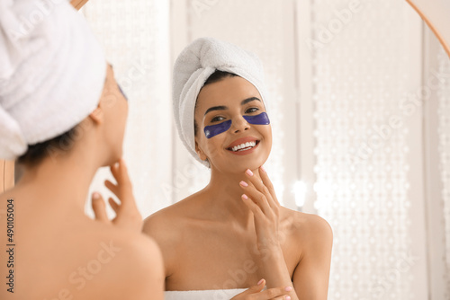 Beautiful young woman with under eye patches near mirror at home
