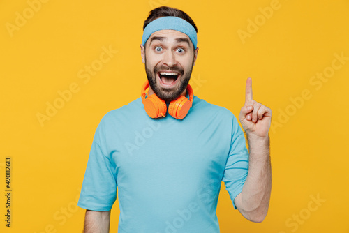 Young fitness trainer instructor sporty man sportsman in headband blue t-shirt spend weekend in home gym hold index finger up with great new idea isolated on yellow background. Workout sport concept.