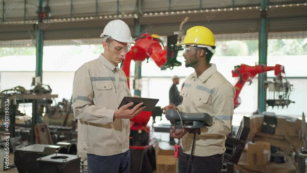 Two engineers man standing in front of robotic arm machine in the factory. Manager worker looking at tablet and read manual how to fix the machine. Maintenance and repairing concept. Brainstorming Two engineers man standing in front of robotic arm machine in the factory. Manager worker looking at tablet and read manual how to fix the machine. Maintenance and repairing concept. Brainstorming