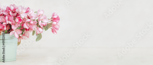 Pink spring flowers in a tiny olive bucket. Pastel beige cozy setup with copy space. Blooming fruit tree branch. Greeting card. Border banner