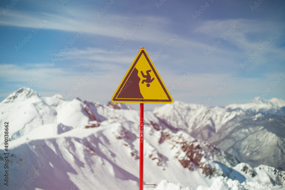 Sign on background of mountains. Sign warning of danger of falling. Triangle on stick. Symbol on yellow background. Carefully abrupt break. Sign on background of mountains. Sign warning of danger of falling. Triangle on stick. Symbol on yellow background. Carefully abrupt break.