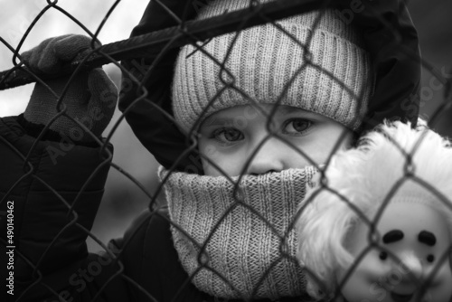 Fotografie, Obraz A little refugee girl with a sad look behind a metal fence