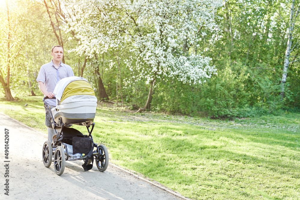 A man with a baby stroller walks through a summer park against the background of blooming apple trees. A child in a white stroller next to his father. A calm atmosphere in nature. High quality photo