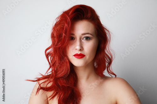 Headshot of stunning young woman with red lips looking at camera on white wall.