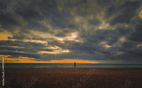 View of dramatic clouds at English seaside at twilight; distant lonely figure standing on gravel beach by water edge