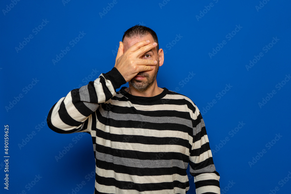 Portrait of a scared man covering his face with palm and looking at camera through fingers isolated on a blue background. Portrait of a scared man covering his face with palm and looking at camera through fingers isolated on a blue background.