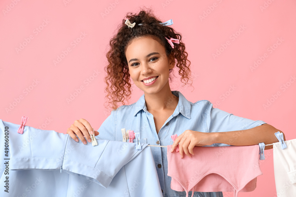 Young African-American woman with hanging laundry and plastic clothespins on pink background Young African-American woman with hanging laundry and plastic clothespins on pink background
