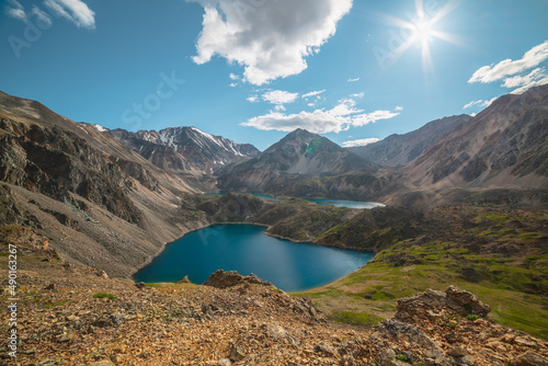 Scenic view to beautiful group of blue mountain lakes at various heights in bright sun. Three blue alpine lakes at different highs among sunlit green hills and large mountains under sun in cloudy sky.